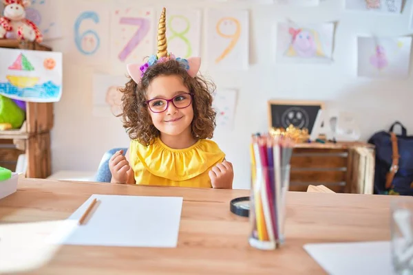 漂亮的幼儿戴着眼镜 骑着独角兽日记坐在幼儿园的课桌上 高兴而兴奋地举起双臂做获奖者的手势 微笑着 尖叫着要成功 庆祝概念 — 图库照片