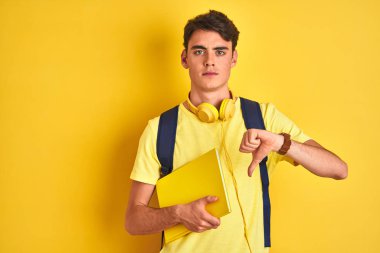 Kulaklık takan ve sırt çantası olan genç bir çocuk izole edilmiş arka planda kitap okuyor. Kızgın yüzlü, negatif işaretli, baş parmakları aşağıda, reddedilme kavramından hoşlanmadığını gösteriyor.