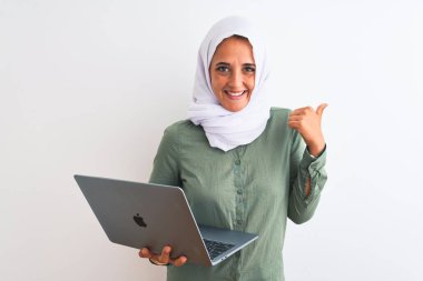 Genç Arap iş kadını tesettür takıyor, bilgisayarla izole edilmiş arka planda işaret ediyor ve gülümseyen bir gülümsemeyle baş parmağıyla gösteriyor.