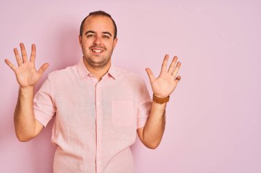 Zarif bir gömlek giyen genç adam pembe arka planda dikilip dokuz numarayı gösterirken kendine güvenen ve mutlu bir şekilde gülümsüyor..