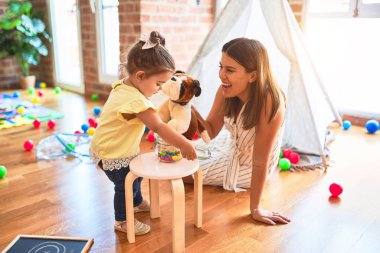 Young beautiful teacher playing with dog doll and toddler holding jar of chocolate balls at kindergarten clipart