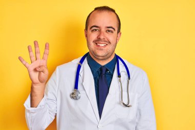 Paltolu ve steteskoplu genç doktor, izole edilmiş sarı arka planda duruyor. Dört numarayı işaret ediyor ve gülümsüyor. Kendinden emin ve mutlu..