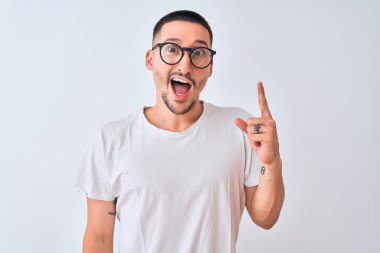 Gözlük takan ve izole edilmiş arka planda duran yakışıklı genç adam başarılı bir fikirle işaret ediyor. Heyecanlı ve mutlu. Bir numara..