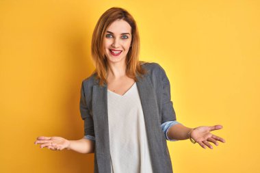Kızıl saçlı beyaz iş kadını sarı arka planda mutlu bir şekilde gülümseyerek dostça karşılanıyor, olumlu ve kendinden emin bir şekilde selamlanıyor.