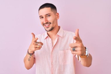 Pembe gömlekli genç adam izole edilmiş arka planda durup mutlu ve komik yüzlü parmakları kameraya doğrultuyor. İyi enerji ve titreşimler.