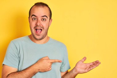 Yeşil günlük tişört giyen genç adam, izole edilmiş sarı arka planda durup hayrete düşmüş ve kameraya gülümserken eliyle ve parmağıyla işaret ediyordu..