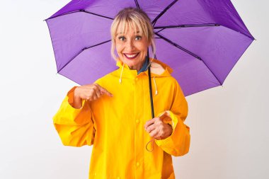 İzole edilmiş beyaz arka planda, yağmurluk ve mor şemsiye giyen orta yaşlı bir kadın kendini işaret eden sürpriz bir yüzle.