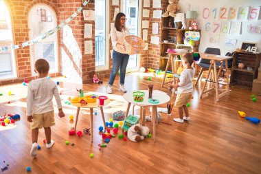 Anaokulunda bir sürü oyuncağın etrafında top ve hasır sepet kullanarak basketbol oynayan güzel öğretmenler ve yeni yürümeye başlayan çocuklar.
