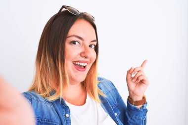 İzole edilmiş beyaz arka planda duran kot gömlek giyen genç ve güzel bir kadın mutlu bir şekilde elini ve parmağını kenarı işaret ediyor.