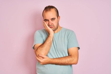Mavi günlük tişört giyen genç bir adam pembe arka planda dikilirken kolları çapraz olan depresyondan sıkılmış ve yorgun görünüyor..