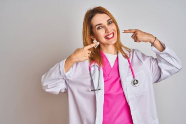 İzole edilmiş arka planda pembe steteskop takan, neşeli gülümseyen ve parmaklarıyla ve ağzıyla işaret eden beyaz saçlı doktor kadın. Diş sağlığı konsepti.