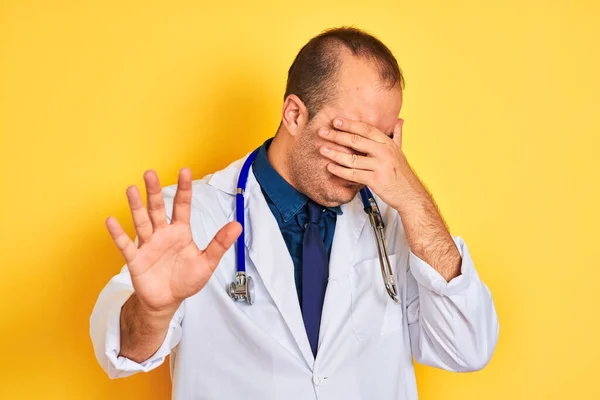 Young doctor man wearing coat and stethoscope standing over isolated yellow background covering eyes with hands and doing stop gesture with sad and fear expression. Embarrassed and negative concept.