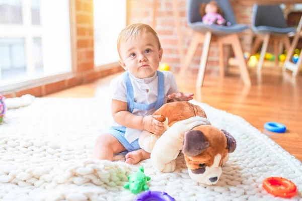 Güzel çocuk, anaokulunda battaniyeye oturmuş oyuncak köpekle oynuyor.