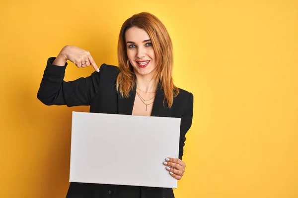 Redhead business caucasian woman holding banner over yellow isolated background with surprise face pointing finger to himself