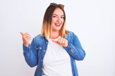 İzole edilmiş beyaz arka planda duran kot gömlekli güzel bir kadın. Arka tarafı işaret ediyor. El ve baş parmakları havada, kendine güvenen.
