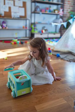 Anaokulunda bir sürü oyuncağın etrafında arabayla oynayan sevimli sarışın çocuk.