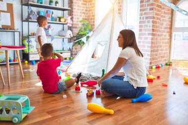 Young beautiful teacher and toddlers playing around lots of toys at kindergarten clipart