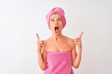 İzole edilmiş beyaz arka plan üzerinde duran orta yaşlı kadın şaşırmış ve şaşırmış bir şekilde yukarı bakıyor ve parmaklarıyla işaret ediyor ve kollarını kaldırıyordu..