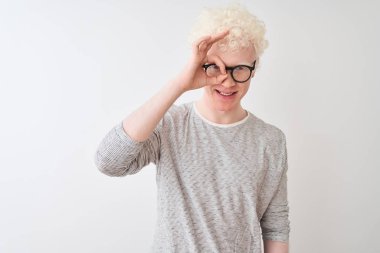 Genç albino sarışın adam çizgili tişört ve gözlüklerle izole edilmiş beyaz arkaplanda el hareketleriyle gülümsüyor ve mutlu yüzle parmaklarının arasından bakıyor..
