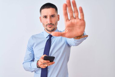 Genç yakışıklı iş adamı izole edilmiş arka planda akıllı telefon kullanıyor. Açık elle dur işareti yapıyor. Ciddi ve kendinden emin bir ifadeyle, savunma hareketi yapıyor.