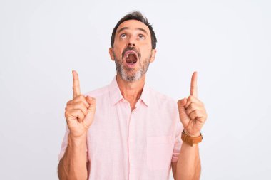Orta yaşlı, pembe gömlekli yakışıklı bir adam izole edilmiş beyaz arka planda duruyor. Şaşırmış ve şaşırmış bir şekilde yukarı bakıyor ve parmaklarıyla işaret ediyor ve kollarını kaldırıyor..