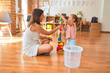 Güzel öğretmen ve sarışın küçük kız kreşteki plastik blokları kullanarak kule inşa ediyorlar.