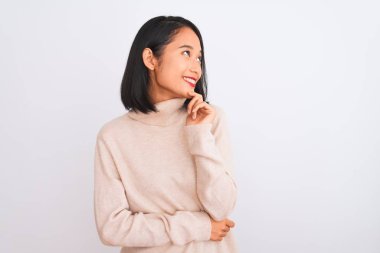 Balıkçı yaka kazak giyen genç Çinli bir kadın izole edilmiş beyaz arka plan üzerinde duruyor ve elleri çenesinde soru sormayı, düşünceli ifadeyi düşünüyor. Gülen ve düşünceli bir yüz. Şüphe kavramı.