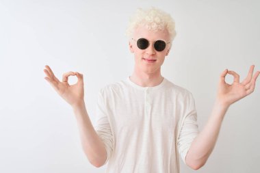 Ayrı beyaz arka planda tişört ve güneş gözlüğü takan genç albino sarışın adam rahatla ve gözleri kapalı meditasyon yaparken parmaklarınla gülümse. Yoga konsepti.