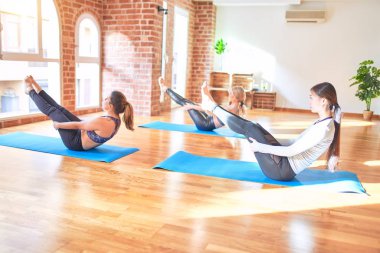 Gülümseyen, yoga yapan genç ve güzel bir kadın grubu. Oturmuş spor salonunda tekne pozu veriyor.
