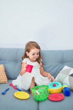 Sevimli sarışın bebek kanepede oturuyor evde yemek ve çatal bıçakla yemek pişiriyor.