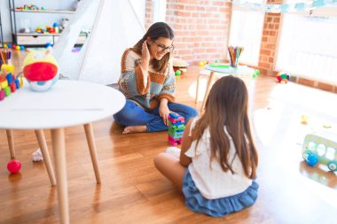 Beautiful teacher and toddler playing with wooden building blocks around lots of toys at kindergarten clipart