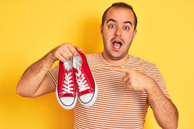 İzole edilmiş sarı arka planda kırmızı spor ayakkabıları tutan genç adam mutlu bir şekilde eli ve parmağı ile işaret ediyor.