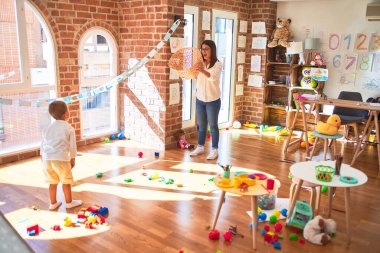 Beautiful teacher and toddler playing basketball using ball and wicker basket around lots of toys at kindergarten clipart