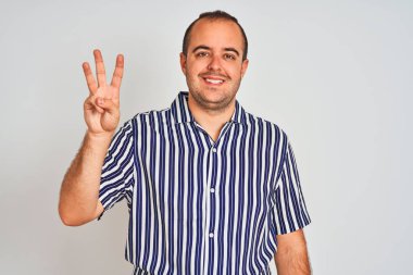 İzole edilmiş beyaz arka planda duran mavi çizgili tişörtlü genç adam üç numaralı parmaklarıyla gülümseyerek kendinden emin ve mutlu bir şekilde duruyor..