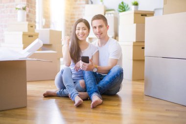 Güzel bir çift, yeni evlerinde karton kutuların etrafında akıllı telefonlar kullanıyor ve gülümseyen bir gülümsemeyle baş parmaklarıyla onları gösteriyorlar.