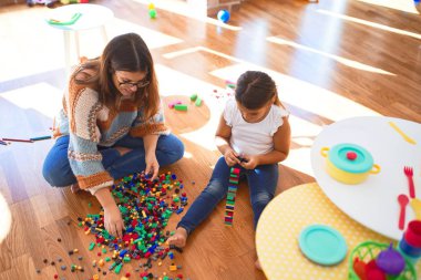 Beautiful teacher and toddler playing with building blocks toy around lots of toys at kindergarten clipart