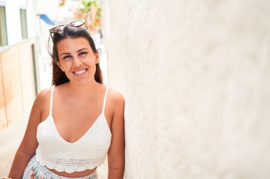 Güzel kız beyaz duvara yaslanıyor, genç arkadaş canlısı kadın mutlu bir yaz gününde gülümsüyor.