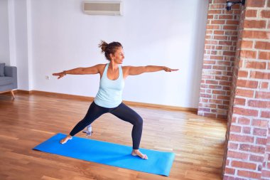 Orta yaşlı, güzel bir sporcu mindere basıyor. Beden eğitiminde yoga yapıyor, savaşçı pozu veriyor.