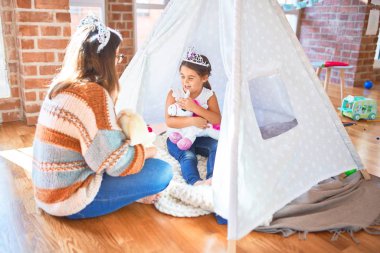 Beautiful teacher and toddler wearing princess crown playing with unicorn doll inside tipi around lots of toys at kindergarten clipart
