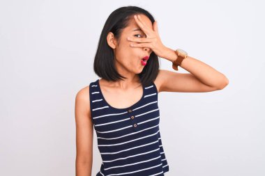 Çizgili tişört giyen genç Çinli bir kadın izole edilmiş beyaz arka planda dikiliyor. Şok olmuş bir şekilde yüzünü ve gözlerini kapıyor. Utanmış bir ifadeyle parmaklarının arasından bakıyor..