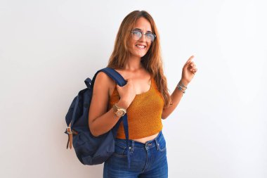 Genç kızıl saçlı, gözlüklü ve sırt çantalı bir bayan izole edilmiş arka planda mutlu bir şekilde el ve parmak uçlarını işaret ediyor.
