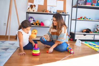 Beautiful teacher and toddler building pyramid using hoops around lots of toys at kindergarten clipart