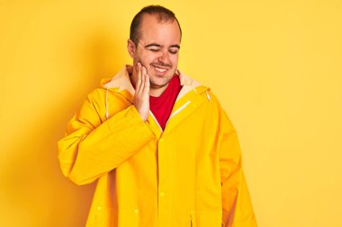 İzole edilmiş sarı arka planda duran yağmurluk giymiş genç bir adam diş ağrısı ya da diş ağrısı yüzünden ağız ve acı dolu bir ifadeyle dokunuyor. Dişçi konsepti.