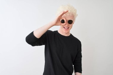 Siyah tişört giyen genç albino adam ve güneş gözlüklü adam izole edilmiş beyaz arka planda dikilip el hareketleriyle gülümsüyor ve mutlu yüzle parmaklarının arasından bakıyor..