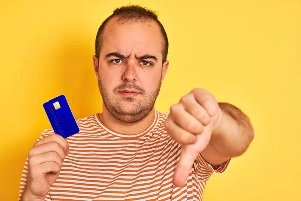 持蓝色信用卡的年轻人站在孤立的黄色背景上 满脸怒容 负面迹象表明不喜欢大拇指朝下 拒绝的概念 — 图库照片