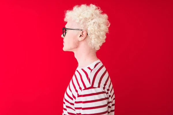 Young albino blond man wearing striped t-shirt and glasses over isolated red background looking to side, relax profile pose with natural face with confident smile.