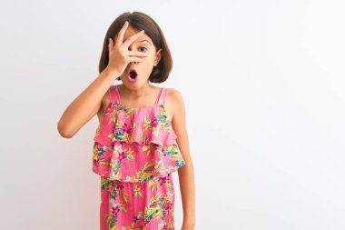 Pembe çiçekli bir elbise giyen genç bir kız çocuğu izole edilmiş beyaz arka planda duruyor. Şok olmuş bir şekilde yüzünü ve gözlerini kapıyor ve utanmış bir ifadeyle parmaklarının arasından bakıyor..