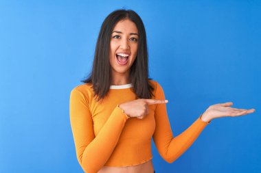 Young beautiful chinese woman wearing orange t-shirt standing over isolated blue background amazed and smiling to the camera while presenting with hand and pointing with finger.