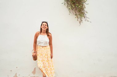 Güzel kız beyaz duvara yaslanıyor, genç arkadaş canlısı kadın mutlu bir yaz gününde gülümsüyor.