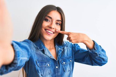 Kot gömlek giyen güzel bir kadın, izole edilmiş beyaz arka planda kamera ile selfie çekiyor. El parmağı ve burunla işaret ediyor, neşeli bir şekilde gülümsüyor. Güzellik konsepti